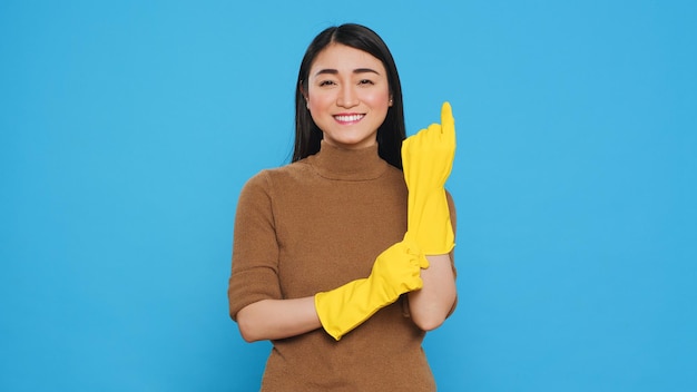 Femme de ménage asiatique souriant à la caméra tout en mettant un gant en caoutchouc après avoir terminé le nettoyage de la maison, debout en studio. Housekepper était habile dans l'art de l'entretien ménager, s'assurant que chaque surface était propre