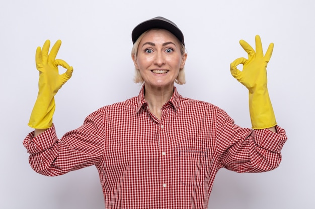 Femme de ménage en chemise à carreaux et casquette portant des gants en caoutchouc regardant la caméra souriant joyeusement montrant un signe ok debout sur fond blanc