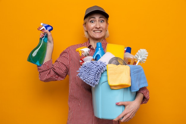 Femme de ménage en chemise à carreaux et casquette tenant une bouteille de produits de nettoyage et un seau avec des outils de nettoyage l'air heureux et surpris