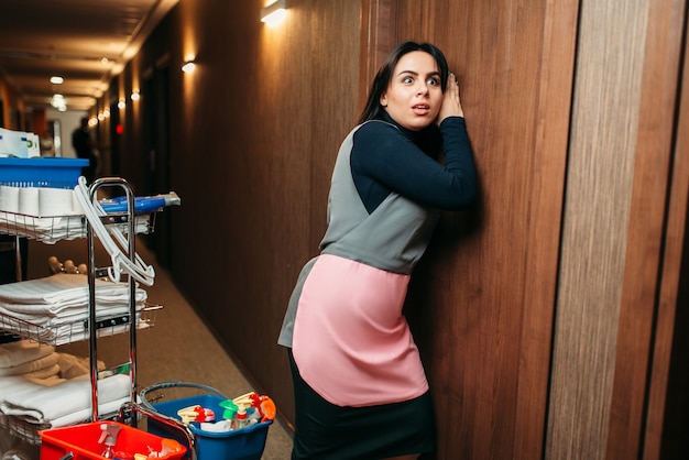 Une femme de ménage curieuse en uniforme surprend à la porte, un chariot avec des détergents, l'intérieur du couloir de l'hôtel. Ménage professionnel, travail de femme de ménage