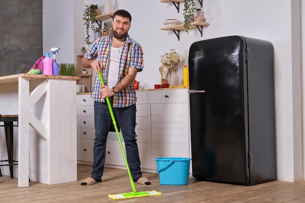 La femme de ménage de mari nettoie la cuisine le jeune homme heureux avec la vadrouille verte lave le sol