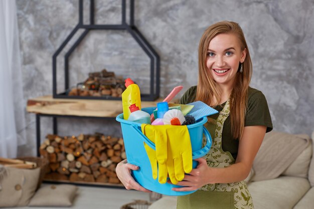 Une femme de ménage se tient dans le salon avec un seau bleu rempli de produits chimiques et d'installations pour ranger dans sa main. Service de nettoyage.