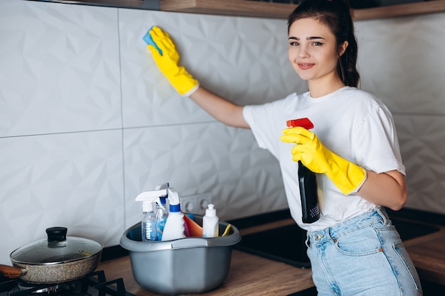 Femme de ménage souriante dans la cuisine de nettoyage de gants jaunes de protection avec des détergents