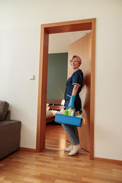 Femme de ménage en uniforme pour le nettoyage de l'appartement contient une boîte avec des détergents et des chiffons et une caméra à la recherche