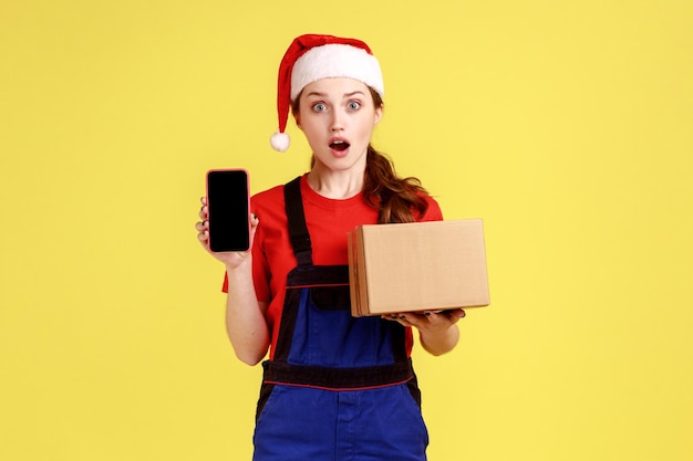 Femme de messagerie étonnée tenant un téléphone portable avec un écran vide pour la promotion et un colis en carton portant une salopette bleue et un chapeau de père noël Prise de vue en studio intérieure isolée sur fond jaune