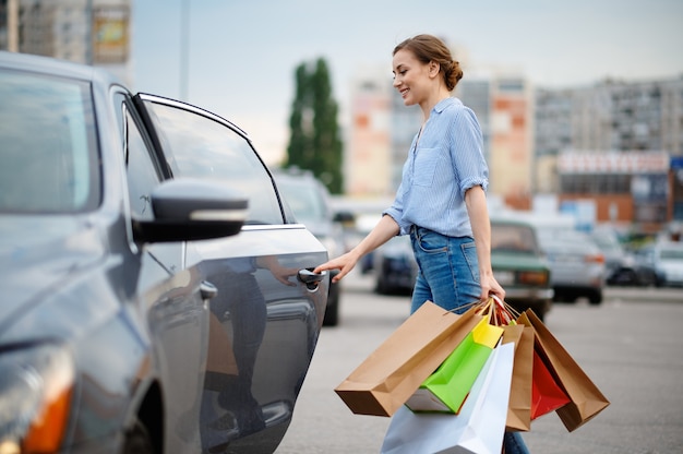 La Femme Met Ses Achats En Voiture Sur Le Parking Du Marché. Client Heureux Transportant Des Achats Du Centre Commercial, Véhicules En Arrière-plan