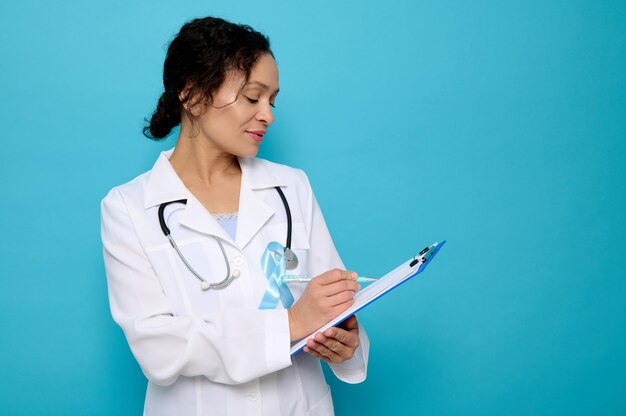 Femme métisse d'âge moyen, femme médecin portant un ruban de satin bleu, couleur symbolique de l'arc sensibilisant à la journée du diabète, maketes sur un presse-papiers, isolée sur fond bleu avec espace de copie