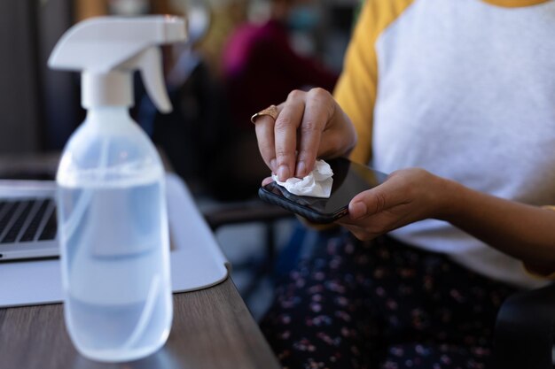 Femme métisse désinfectant son smartphone dans un bureau. hygiène sur le lieu de travail pendant la pandémie du coronavirus covid 19.