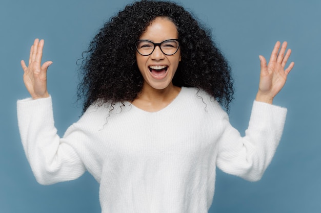 Une femme métisse ravie aux cheveux bouclés lève les mains s'exclame des émotions positives garde la bouche ouverte vêtue d'un pull blanc se dresse sur fond bleu Concept d'expressions faciales