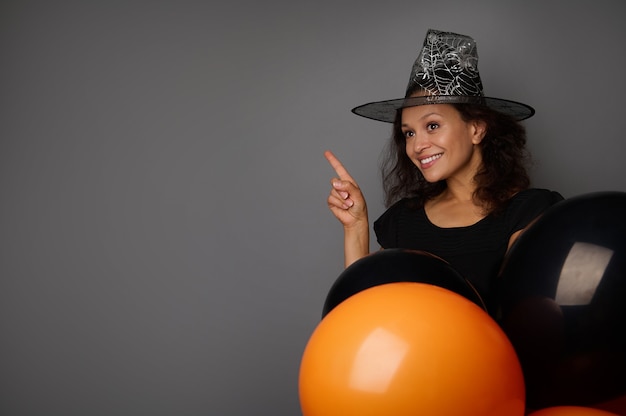 Photo une femme mignonne souriante de race mixte vêtue d'un costume de carnaval de sorcière tient des ballons à air colorés orange et noir, pointe sur un espace de copie sur fond gris. concept d'événement saisonnier traditionnel d'halloween