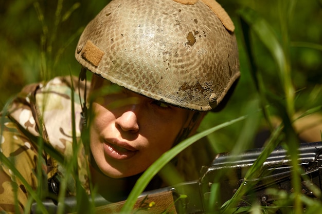 Femme militaire caucasienne en tenue tactique posant pour une photo à la saison estivale Portant un uniforme de camouflage vert et un fusil d'assaut en tenue militaire et une dame de casque regarde à côté