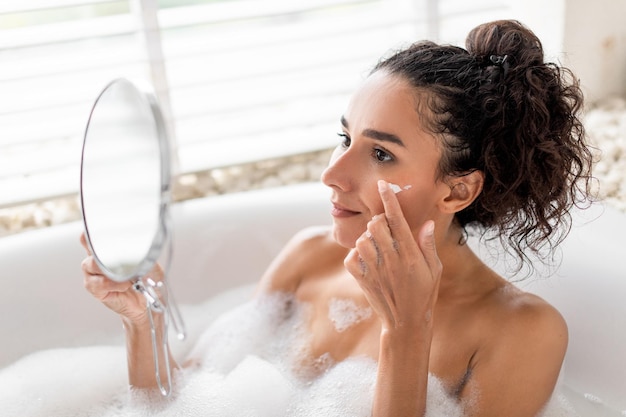 Femme millénaire joyeuse utilisant de la crème pour le visage dans la baignoire regardant dans le miroir à la maison