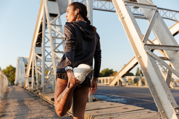 femme mince afro-américaine en tenue de sport utilisant des écouteurs et étirant sa jambe tout en travaillant sur le vieux pont