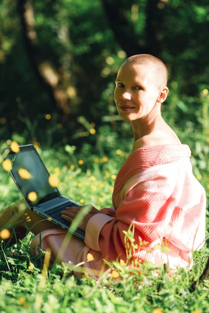 Une femme mince et chauve dans un peignoir avec un ordinateur portable regarde la caméra tout en étant assise sur l'herbe verte à l'extérieur Été printemps saison Concept de magasinage en ligne indépendant satisfait