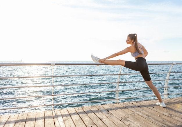 Une femme mince avec un corps parfait pratique l'étirement des jambes avant de s'entraîner sur la plage au lever du soleil