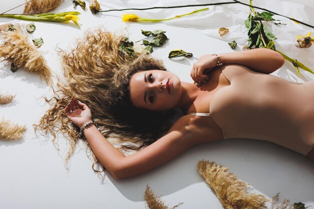 Une femme mince dans une combinaison beige pose à côté de fleurs séchées sur fond blanc isolé Sportive en maillot de bain beige posant à côté de fleurs sur fond blanc