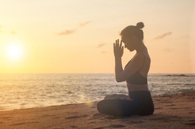 Une femme mince fait du yoga en posture de lotus sur la côte tropicale de la mer ou sur la plage de l'océan en plein air au coucher du soleil