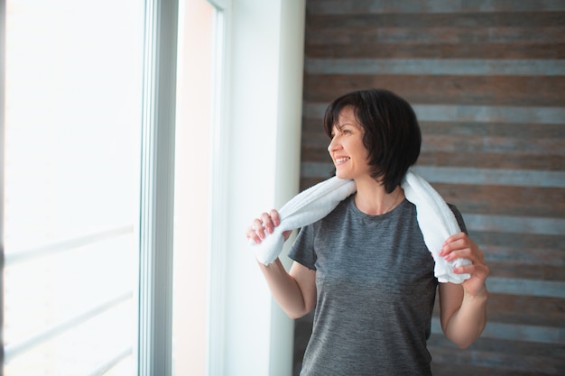 Une femme mince en forme d'adulte fait de l'exercice à la maison. Une femme âgée forte et bien bâtie se tient seule dans la chambre après l'exercice. Tenant une serviette blanche autour du cou. Repose toi et relaxe toi.