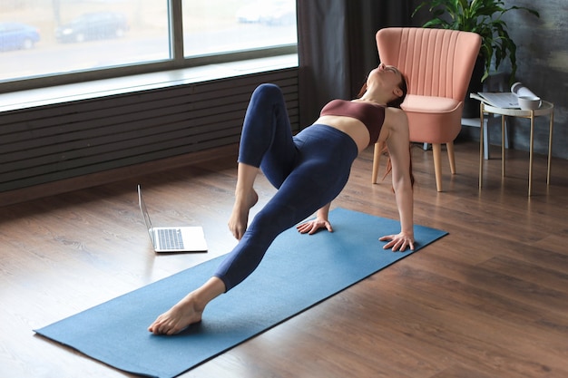 Femme mince sportive pratiquant le yoga avancé devant un ordinateur portable, regardant des tutoriels en ligne. Mode de vie sain.