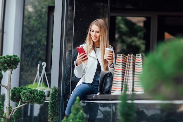Une femme de mode caucasienne avec une tasse de café assise près des vitrines des magasins dans la rue à l'extérieur se repose pendant la pause-café.