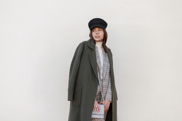 Femme à la mode avec un chapeau dans un manteau vert élégant avec un costume à la mode à carreaux et un sac posant près du mur