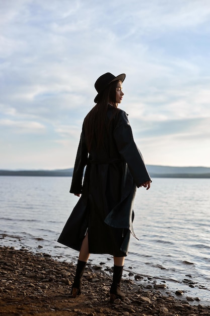 Femme de mode dans le chapeau rond noir et l'imperméable en cuir près de l'hiver de mer de plage. Beau maquillage et rouge à lèvres rouge vif sur les lèvres