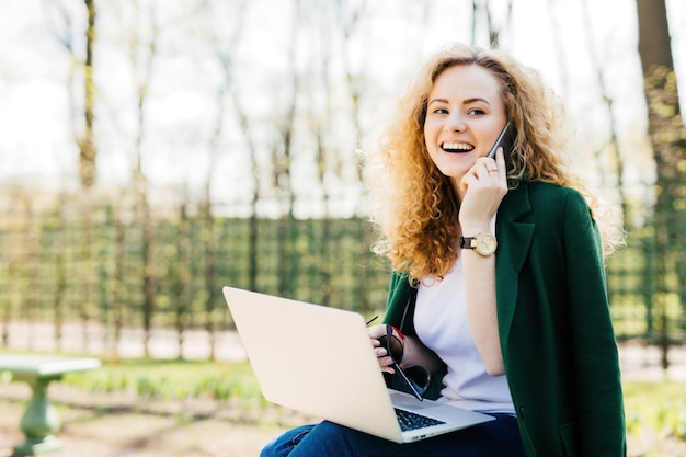 Femme à la mode dans le parc souriant tout en communiquant sur un téléphone portable et à l'aide d'un ordinateur portable