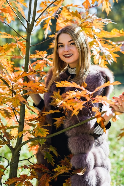 Femme de mode. Jeune fille souriante en manteau de fourrure posin en automne parc avec arbres et lierre