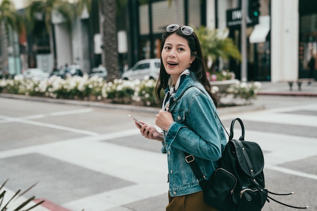 femme de mode marchant joyeusement sur la route, tenant son smartphone et portant un sac à dos noir