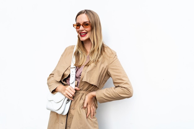 Femme à la mode à la mode élégante portant un sac en cuir trench-coat et des lunettes de soleil posant sur fond blanc automne automne look de mode de rue