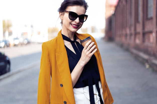 Femme de mode portrait à lunettes de soleil marchant dans la rue. Elle porte une veste jaune, souriant à côté.