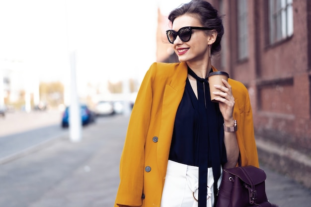 Femme de mode portrait à lunettes de soleil marchant dans la rue. Elle porte une veste jaune, souriant à côté.