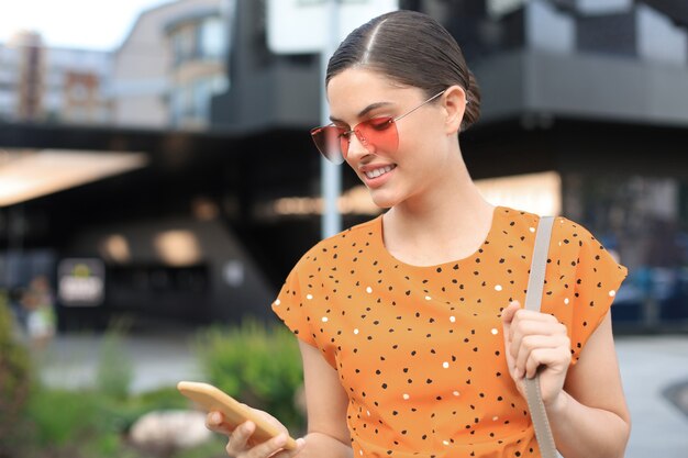 Femme de mode portrait en robe jaune à lunettes de soleil marchant dans la rue et tenant le smartphone dans sa main.