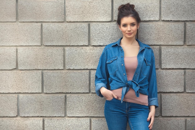 Femme de mode de style de rue dans des vêtements en denim debout près d'un mur de briques vides