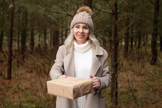 Femme à la mode vêtements chauds tenant une boîte-cadeau dans un emballage en papier Kraft sur fond de nature