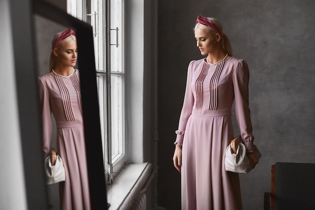 Une femme modèle élégante avec un maquillage parfait vêtue d'une robe de soirée rose en gardant le sac à main et posant dans un intérieur vintage près du miroir