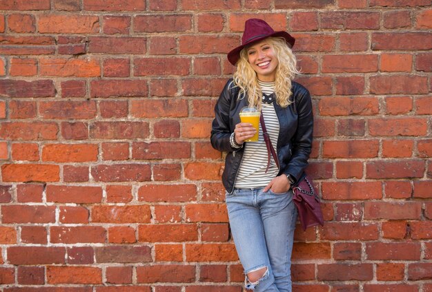 Femme moderne debout près du mur de briques avec du jus d'orange dans les mains