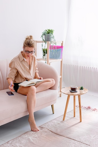 Une femme moderne est assise sur un canapé confortable dans un intérieur lumineux écrit dans un journal