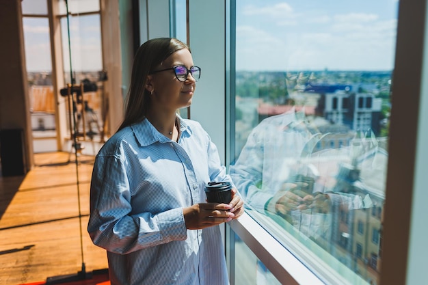 Une femme moderne regarde des vidéos sur un ordinateur portable et boit du café Le concept de distance ou d'apprentissage en ligne Une jeune femme souriante est assise sur une chaise à la table