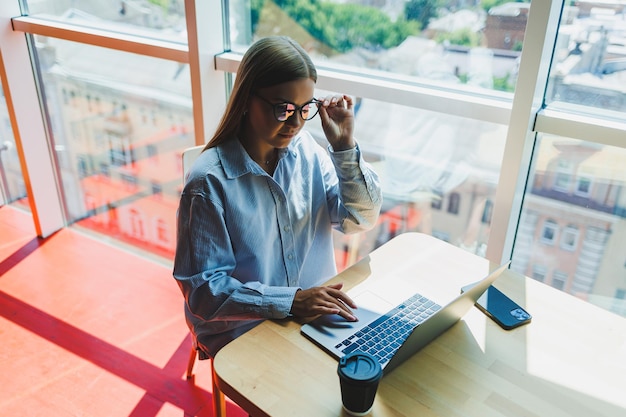 Une femme moderne regarde des vidéos sur un ordinateur portable et boit du café Le concept de distance ou d'apprentissage en ligne Une jeune femme souriante est assise sur une chaise à la table