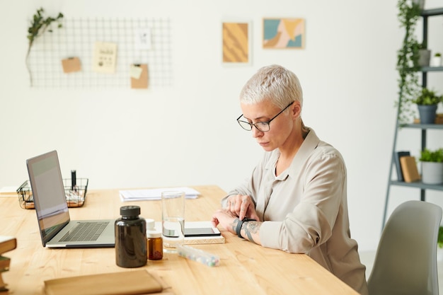 Femme moderne sérieuse aux cheveux blonds vérifiant le temps avant de prendre des vitamines dans le concept de biohacking de bureau