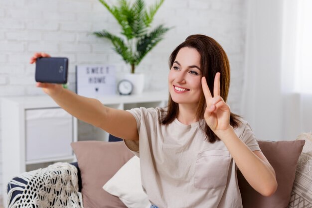 Photo femme montrant un signe de victoire et prenant une photo selfie avec un téléphone intelligent dans un salon confortable