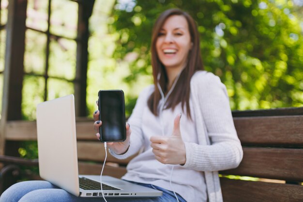 Femme montrant sur téléphone mobile appareil photo avec écran vide vide pour copier l'espace. Femme assise sur un banc travaillant sur un ordinateur portable moderne dans la rue à l'extérieur. Bureau mobile. Concept d'entreprise indépendant.