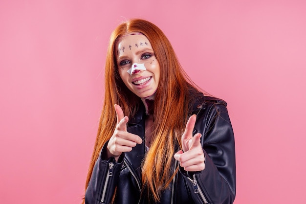 Femme de motard au gingembre aux cheveux roux portant une veste en cuir noire avec une chirurgie de maquillage du sang fracture du nez et du cou pour la fête d'halloween, fond rose studio.