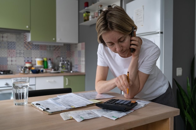 Une femme mûre appelle différentes lignes directes au téléphone pour résoudre des problèmes d'obligations en regardant des factures