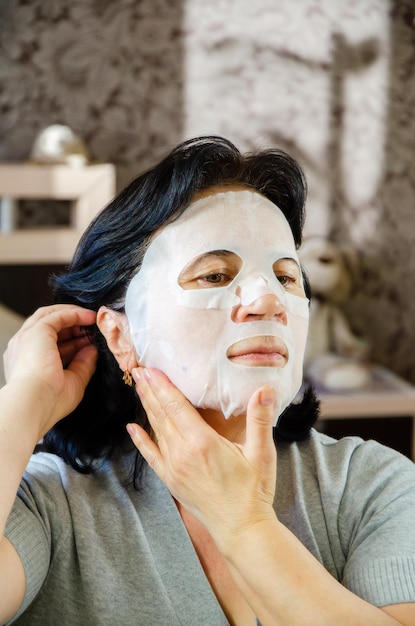 Femme mûre applique un masque cosmétique en tissu sur son visage devant le miroir Soins de la peau pour les personnes âgées