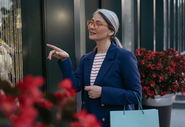 Femme mûre asiatique pensive tenant un sac à provisions en choisissant des vêtements dans le concept de vente du centre commercial