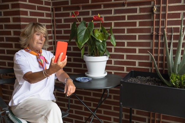 Femme mûre assise sur un balcon avec un téléphone portable prenant un selfie