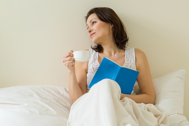 Femme mure au lit, lecture de livre et boire du café