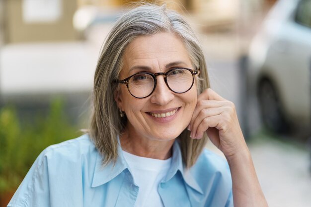 Femme mûre charmante et confiante souriante, penchée la tête sur la main, portant des lunettes et de la merde bleue, assise à l'extérieur avec fond de ville Portrait de femme mûre avec des cheveux gris et une bonne peau